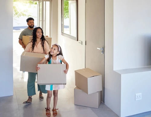 A smiling family carrying moving boxes into a bright new home, ready to unpack and start settling in together. 